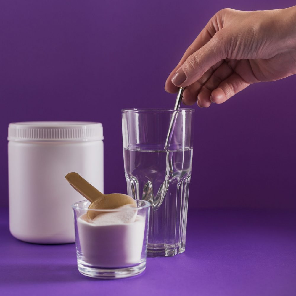 Glass with collagen dissolved in water and collagen protein powd Glass with collagen dissolved in water and collagen protein powder on purple background. Woman's hand holds a spoon. Healthy lifestyle concept.